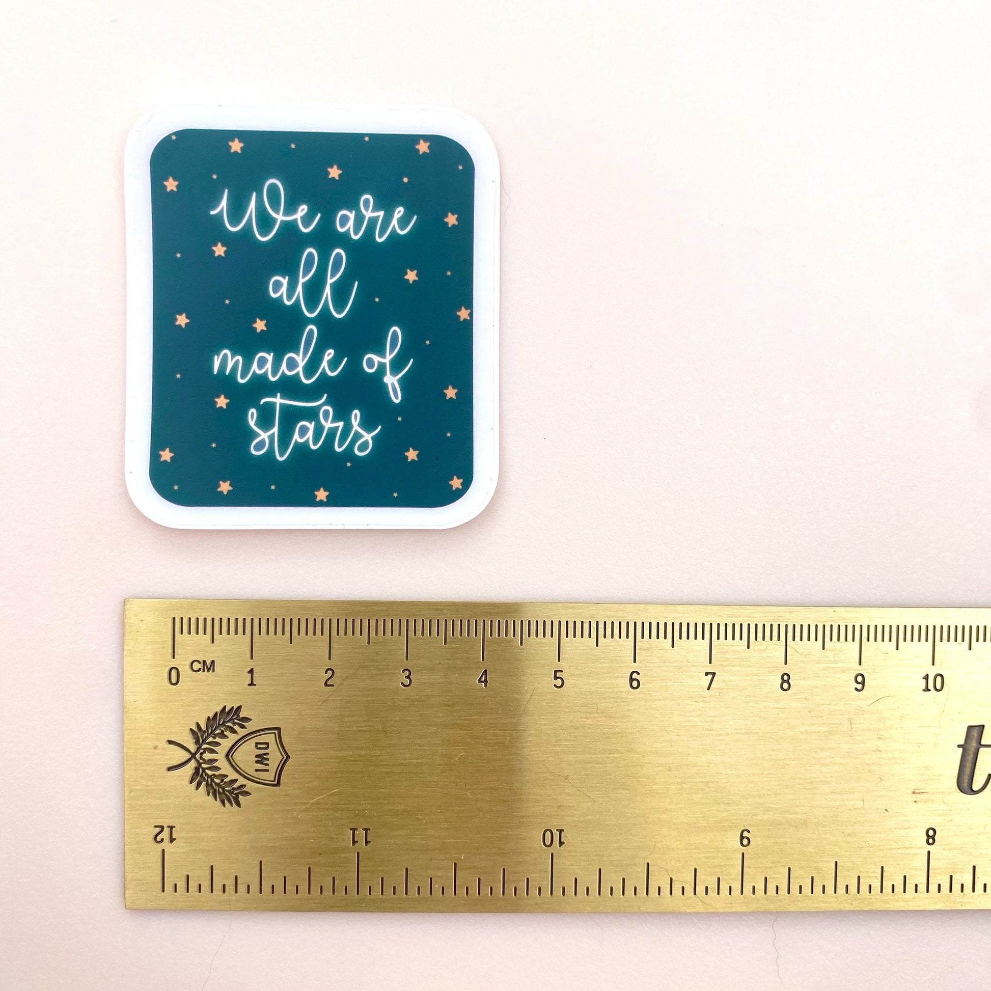 A blue sticker with white glowing text 'We are all made of stars' on a cream background with a gold ruler for scale.