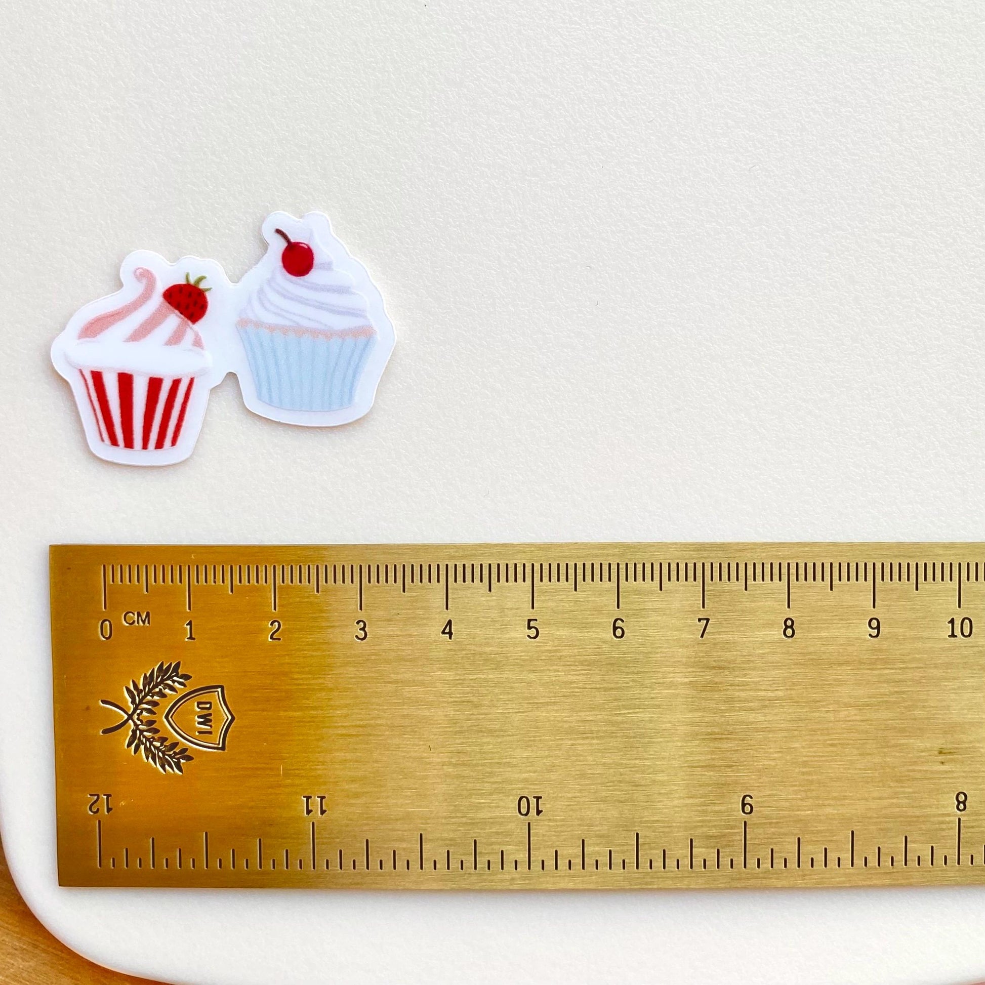 A sticker with two tiny cupcakes in pink and blue against a cream background with a gold ruler for scale.