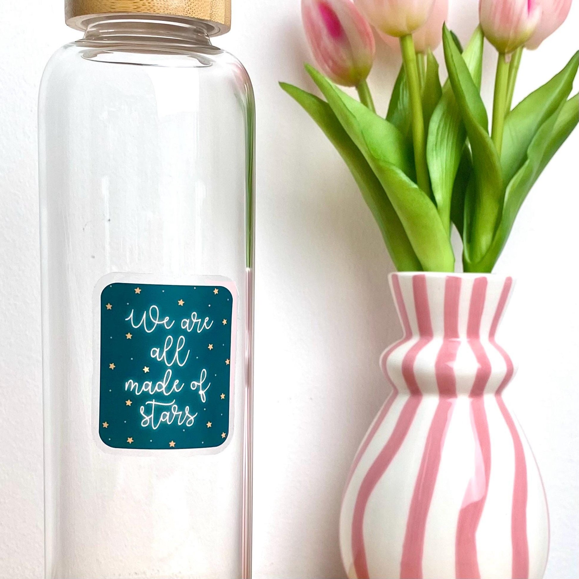 A blue sticker with white glowing text 'We are all made of stars' against a clear glass bottle besides a pink striped vase of tulips.