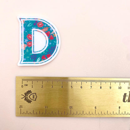Floral letter 'D' sticker next to a ruler on a beige background