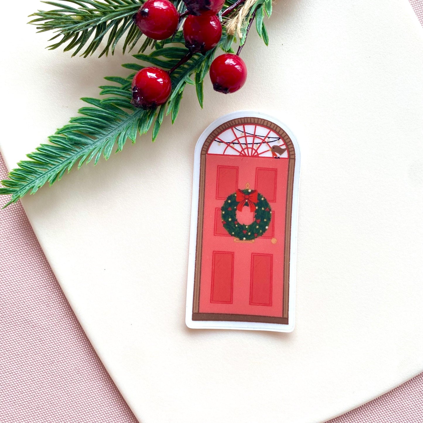 Decorative sticker of a red door with a wreath, surrounded by holly leaves and berries on a light background.