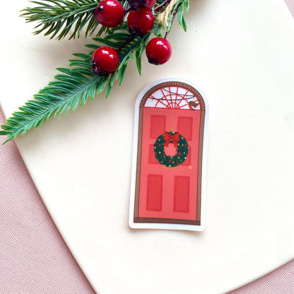 Decorative sticker of a red door with a wreath, surrounded by holly leaves and berries on a light background.