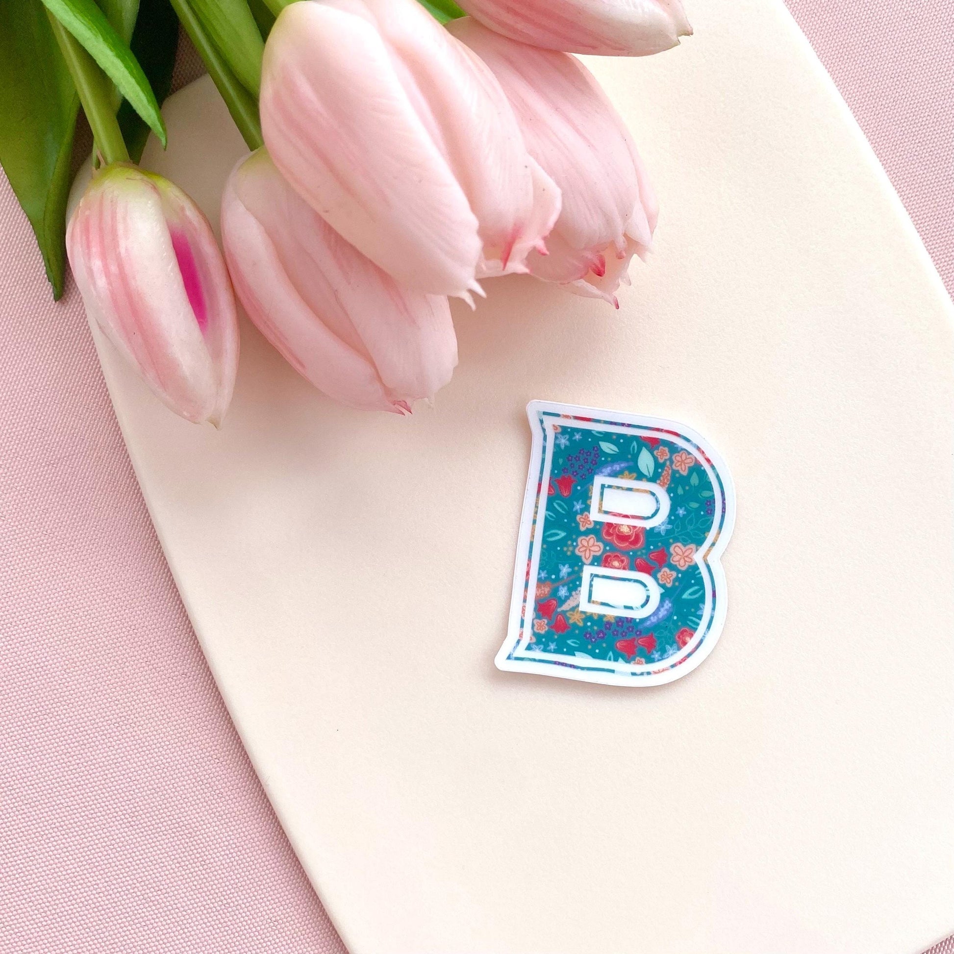 Clear sticker with a teal floral decorated letter 'B' against a cream background with pink tulips.