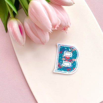 Clear sticker with a teal floral decorated letter 'B' against a cream background with pink tulips.