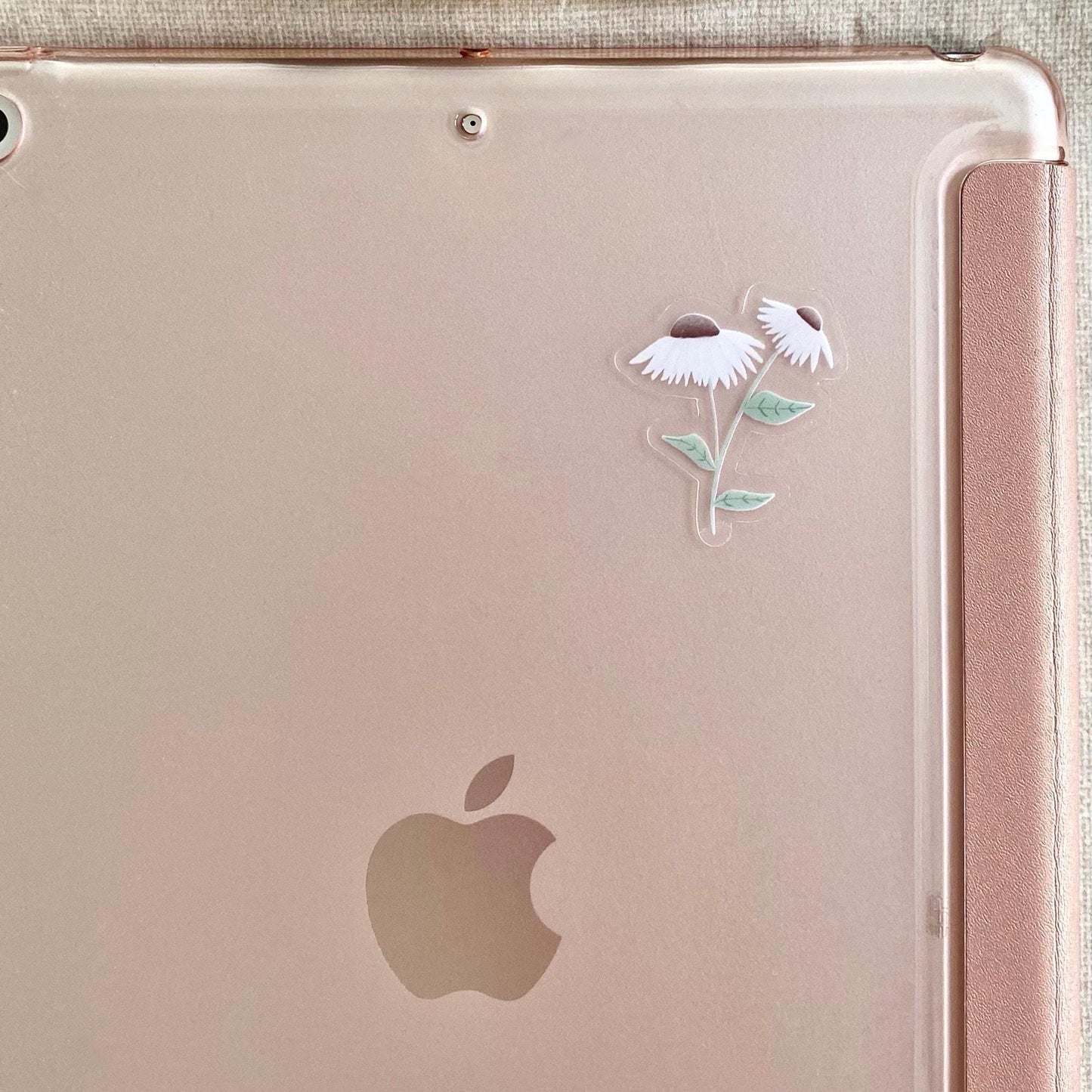 A clear sticker with daisies with white petals and green leaves stuck on the back of a rose gold ipad