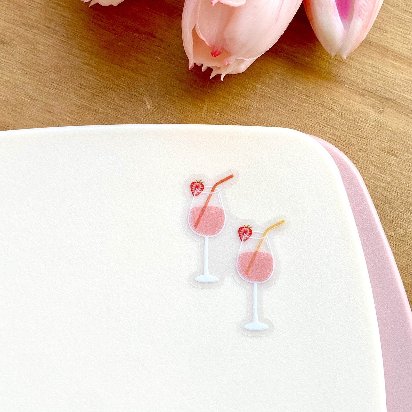 A sticker of two pink drinks in wine glasses with colourful straws and strawberries on a white surface with a wooden background.