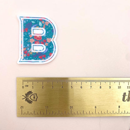 Clear sticker with a teal floral decorated letter 'B' against a cream background with a gold ruler for scale.