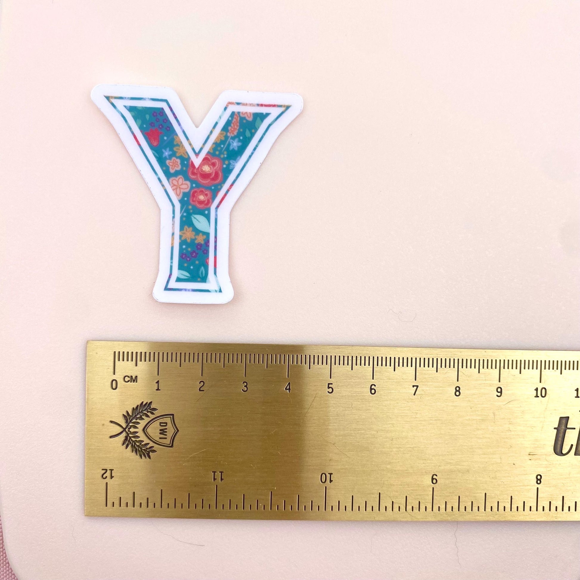 Floral letter 'Y' sticker next to a ruler on a pink background