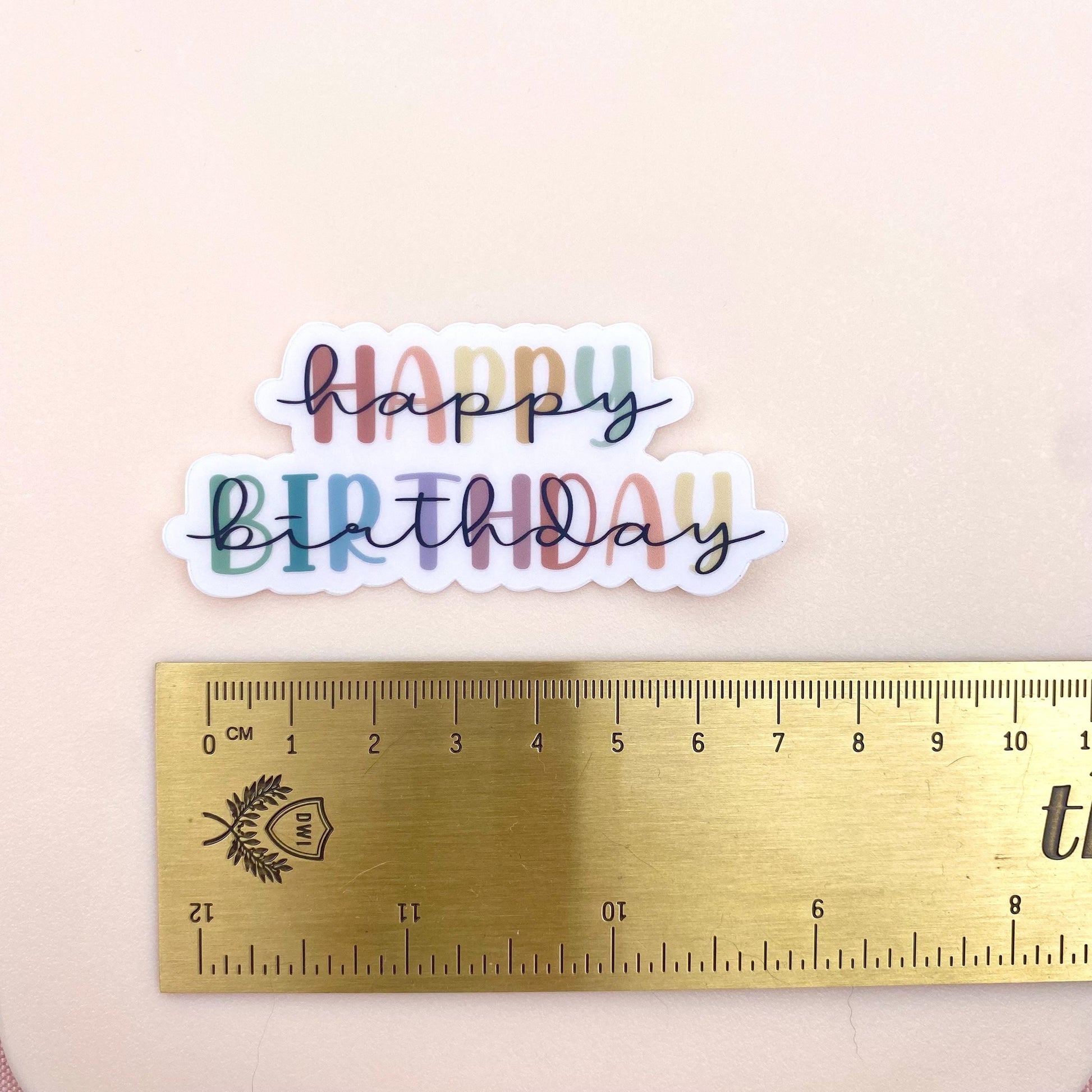 Colourful 'Happy Birthday' sticker with a ruler for scale on a beige background