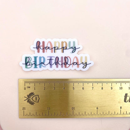 Colourful 'Happy Birthday' sticker with a ruler for scale on a beige background