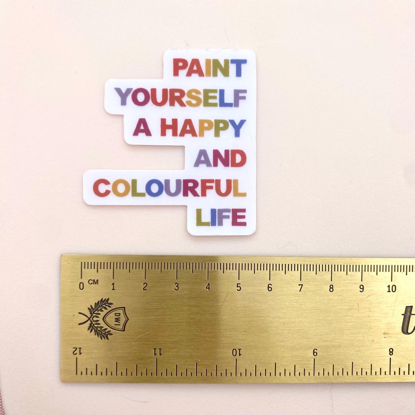 A clear sticker with motivational quote 'Paint yourself a happy and colourful life' on a white background with a gold ruler for scale.