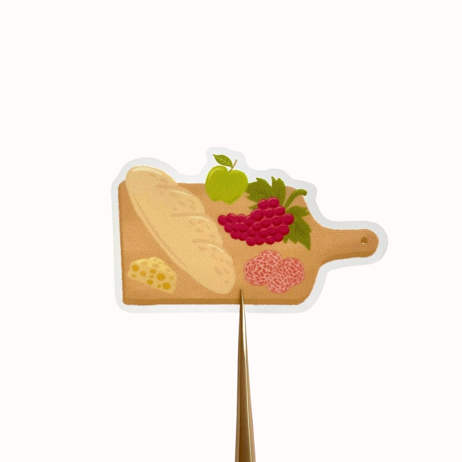 Charcuterie board sticker with a baguette, cheese, grapes, apple and salami, against a cream background