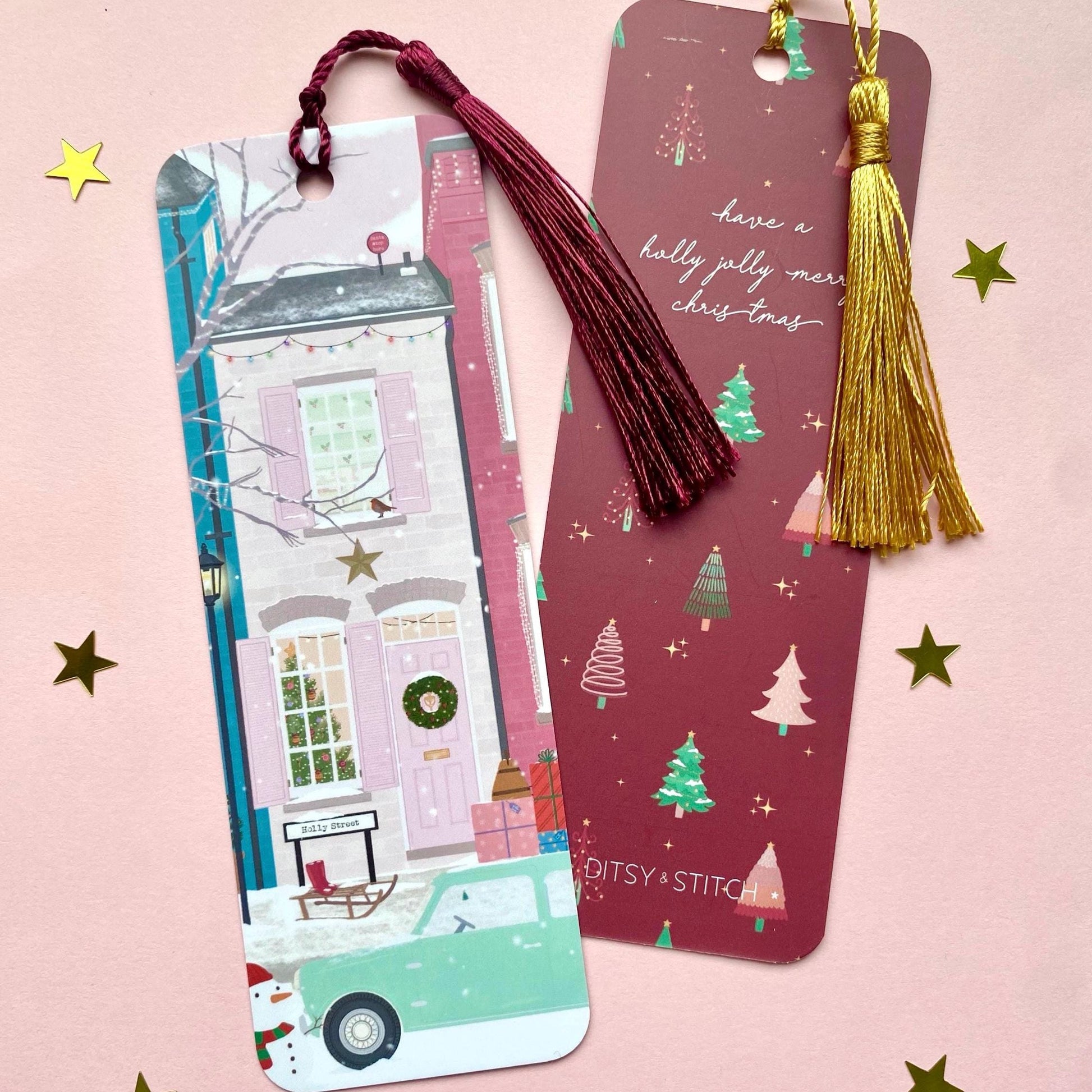 Decorative bookmark depicting a wintery street scene with pink houses with gold tassel on a pink background. Second bookmark showing design on the back with a dark pink Christmas trees pattern and holly jolly quote