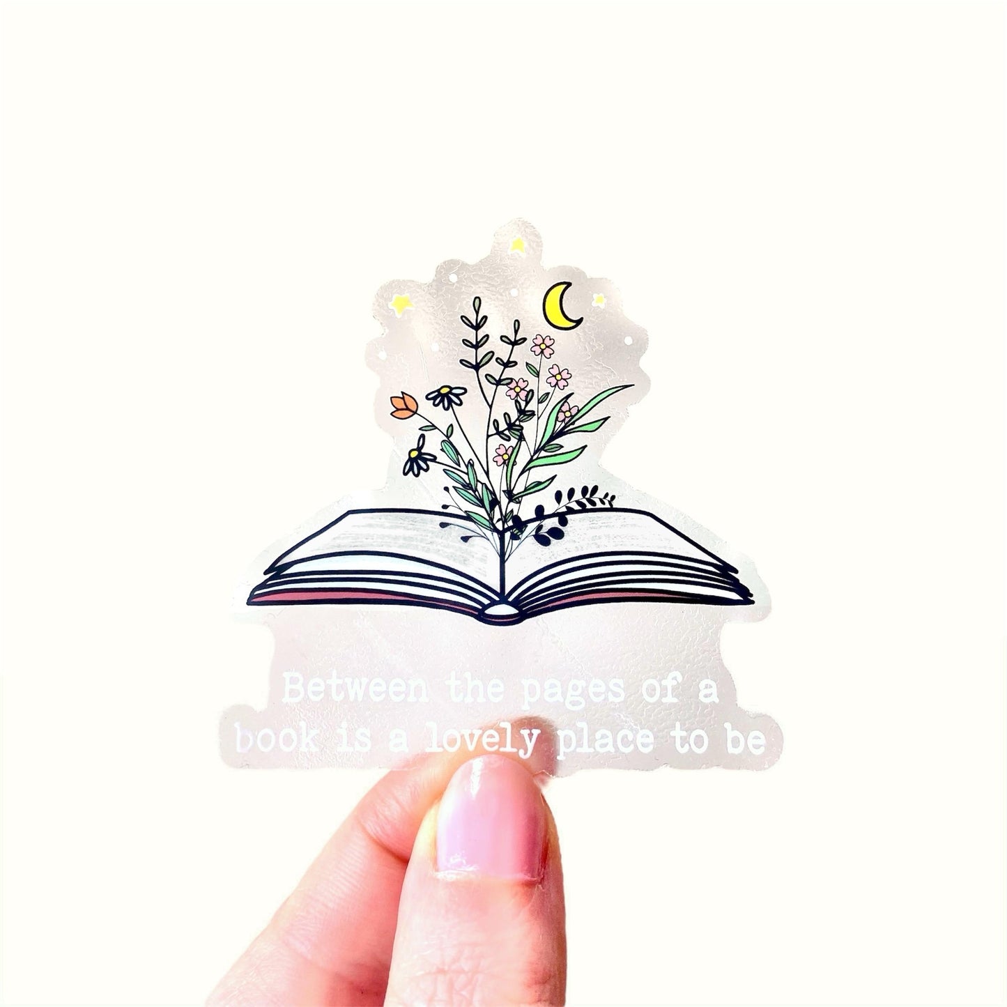 Decorative clear sticker with an open book design and floral elements and a 'Between the pages of a book is a lovely place to be' quote held by a hand.
