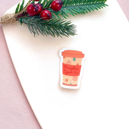 Sticker of a pumpkin spice latte cup on a white surface with autumn foliage.