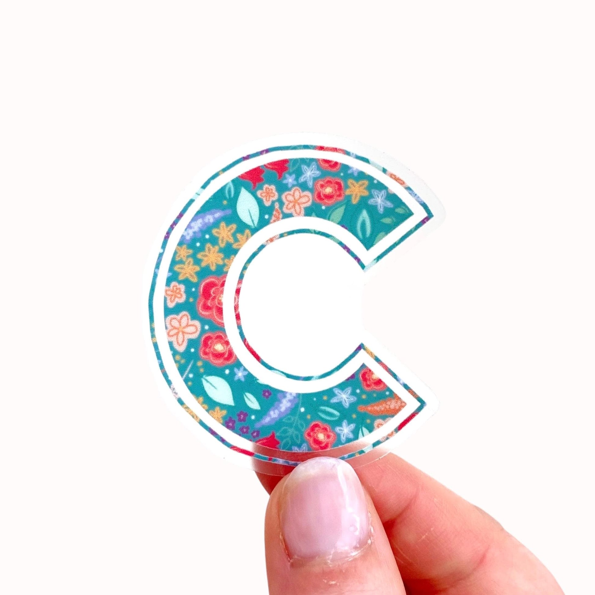 Colorful floral letter 'C' sticker held by a hand on a white background