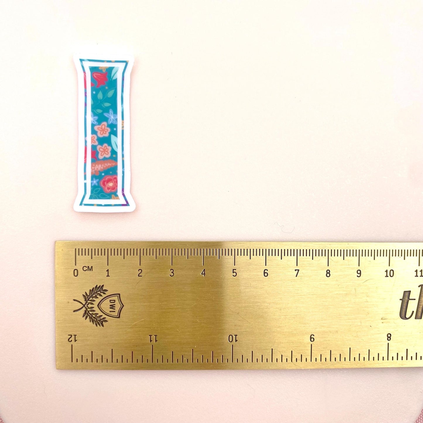 Clear vinyl sticker with a teal floral patterned letter 'I' shown on a white background with a gold ruler for scale