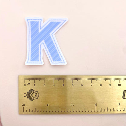 Blue striped letter 'K' transparent sticker show with a gold ruler for scale against a white background