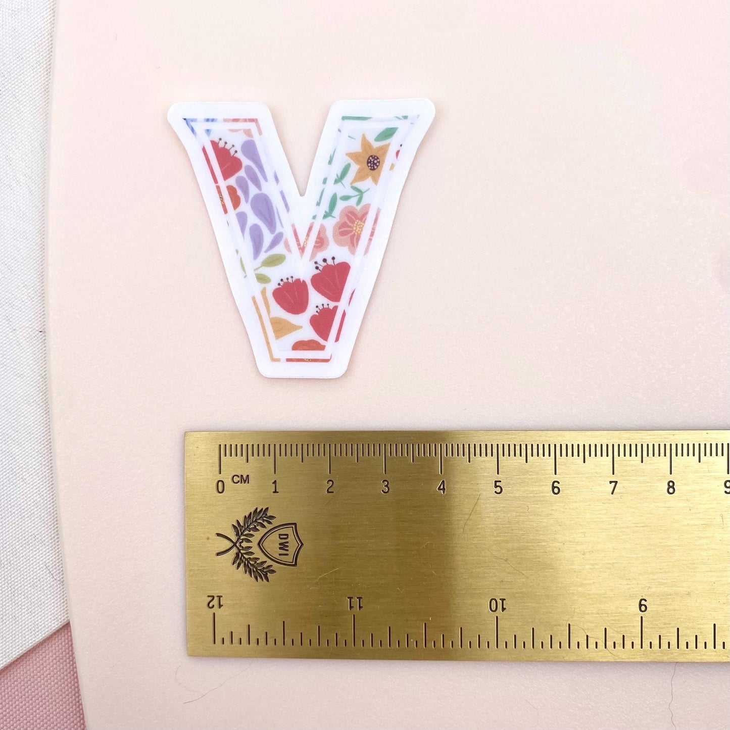 A floral letter 'V' sticker against a white background with a gold ruler for scale.
