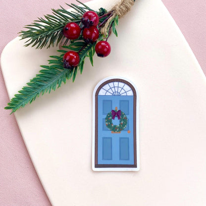 A sticker with a blue door decorated for Christmas against a cream background with festive foliage.