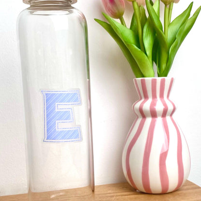 Clear glass bottle with a blue letter 'E' transparent sticker next to a striped pink and white vase with tulips.