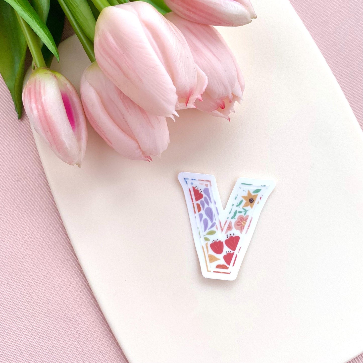 A floral letter 'V' sticker against a white background with pink tulips in the background
