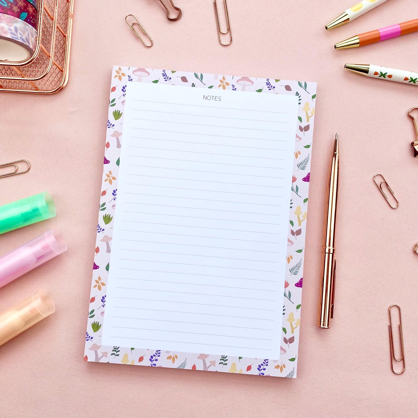 A lined notepad with a beige border patterned with autumn leaves and mushrooms, on a pink background and surrounded by rose gold stationery items