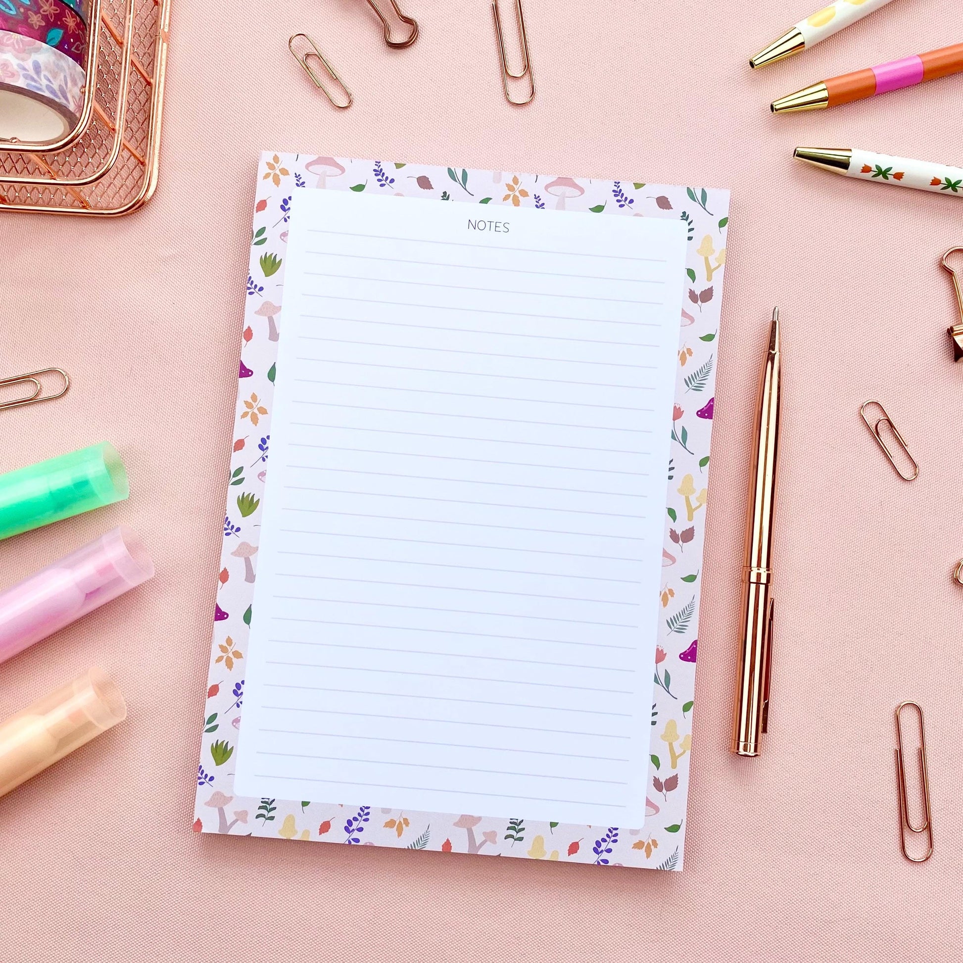 A lined notepad with a beige border patterned with autumn leaves and mushrooms, on a pink background and surrounded by rose gold stationery items