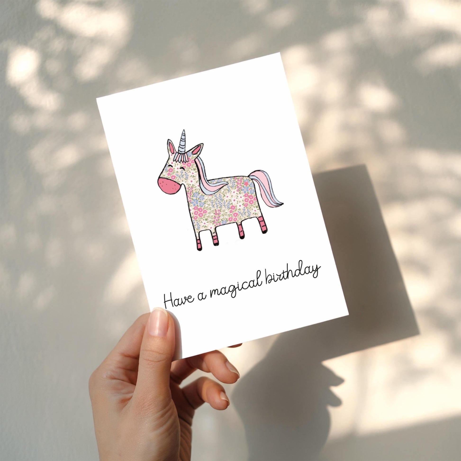Hand holding a card with a unicorn illustration and 'Have a magical birthday' text.