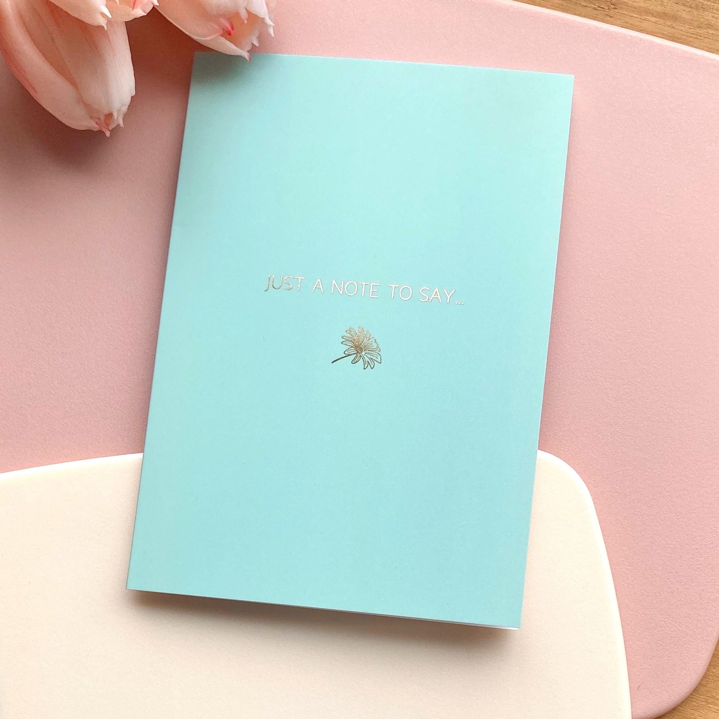 Light blue card with 'Just a Note to Say' gold foiled text and dandelion illustration on a pink surface.
