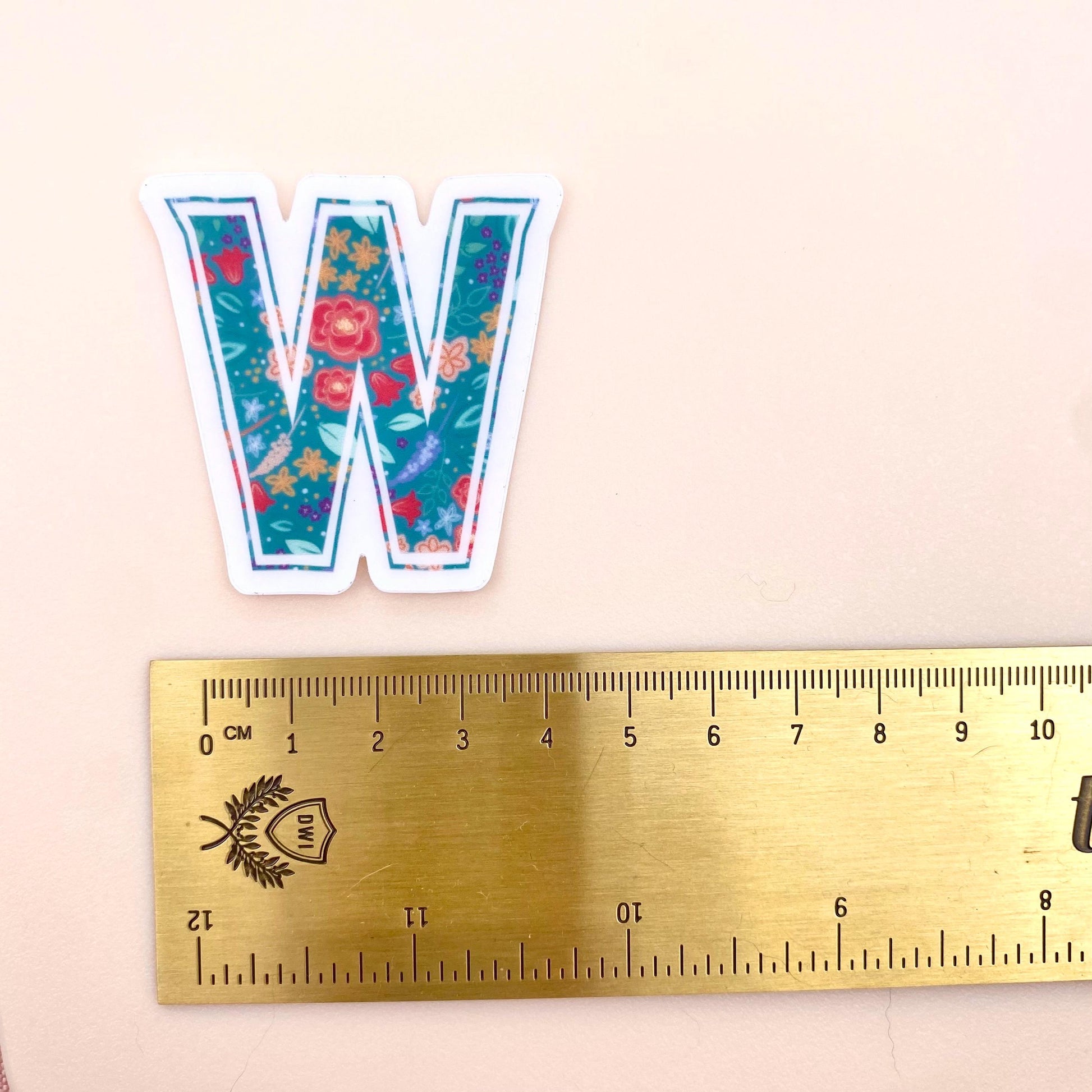 Floral-patterned letter 'W' sticker next to a ruler on a beige background