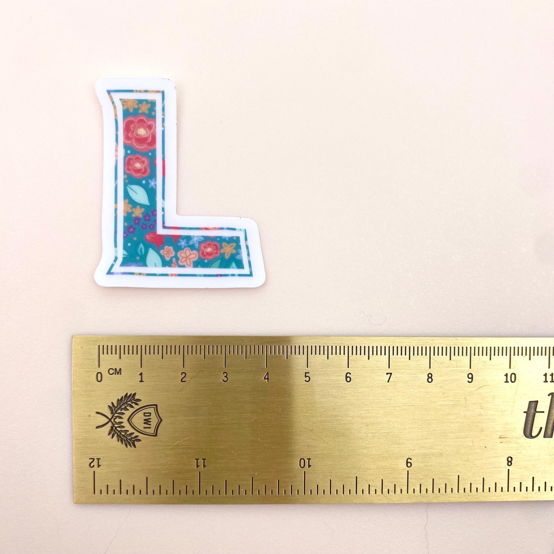 Floral letter 'L' sticker next to a ruler on a beige background