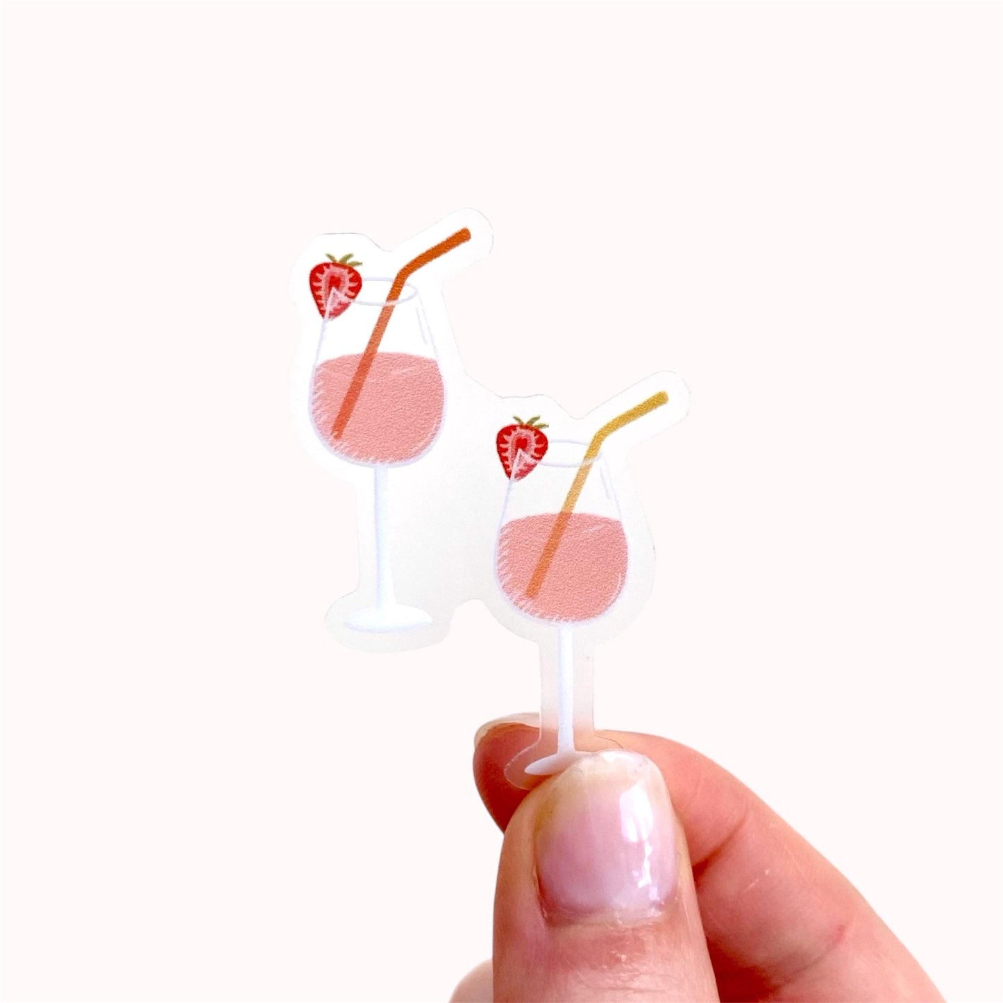 Hand holding a sticker of two pink drinks in wine glasses with colourful straws and strawberries against a white surface.