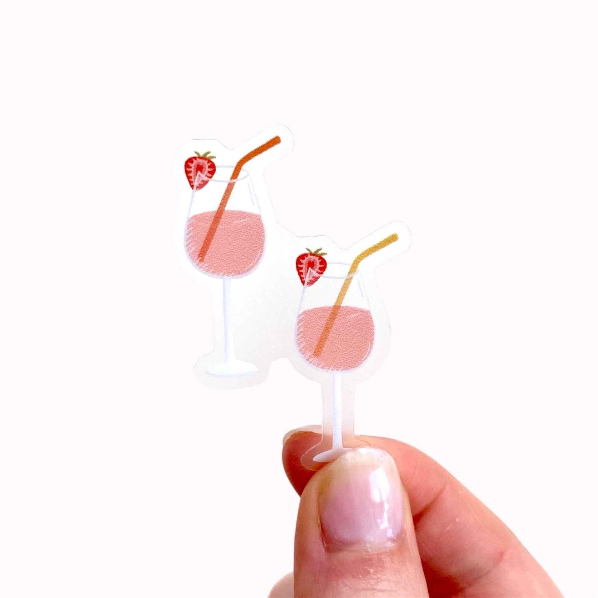 Hand holding a sticker of two pink drinks in wine glasses with colourful straws and strawberries against a white surface.