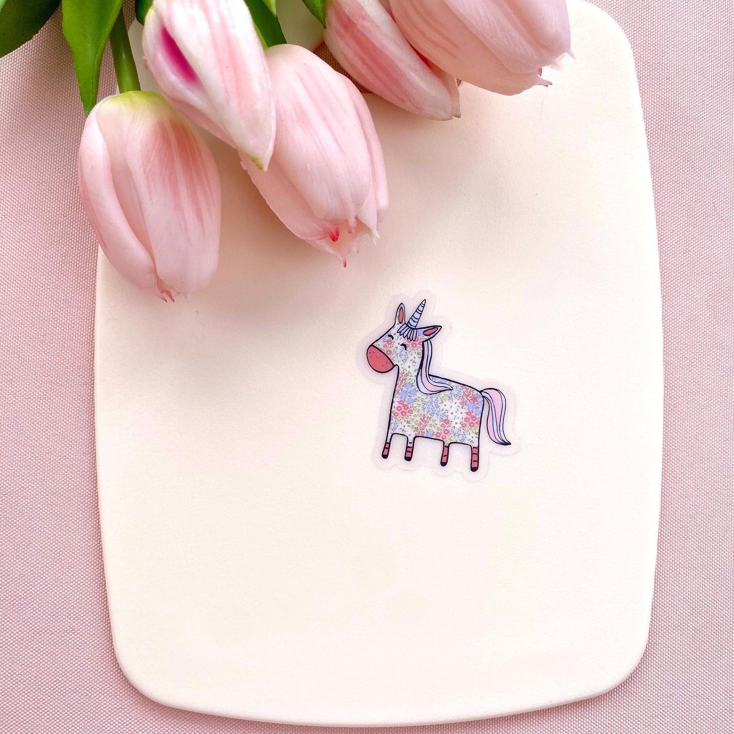 Floral patterned pink and blue unicorn on a transparent vinyl sticker against a white back ground with pink tulips