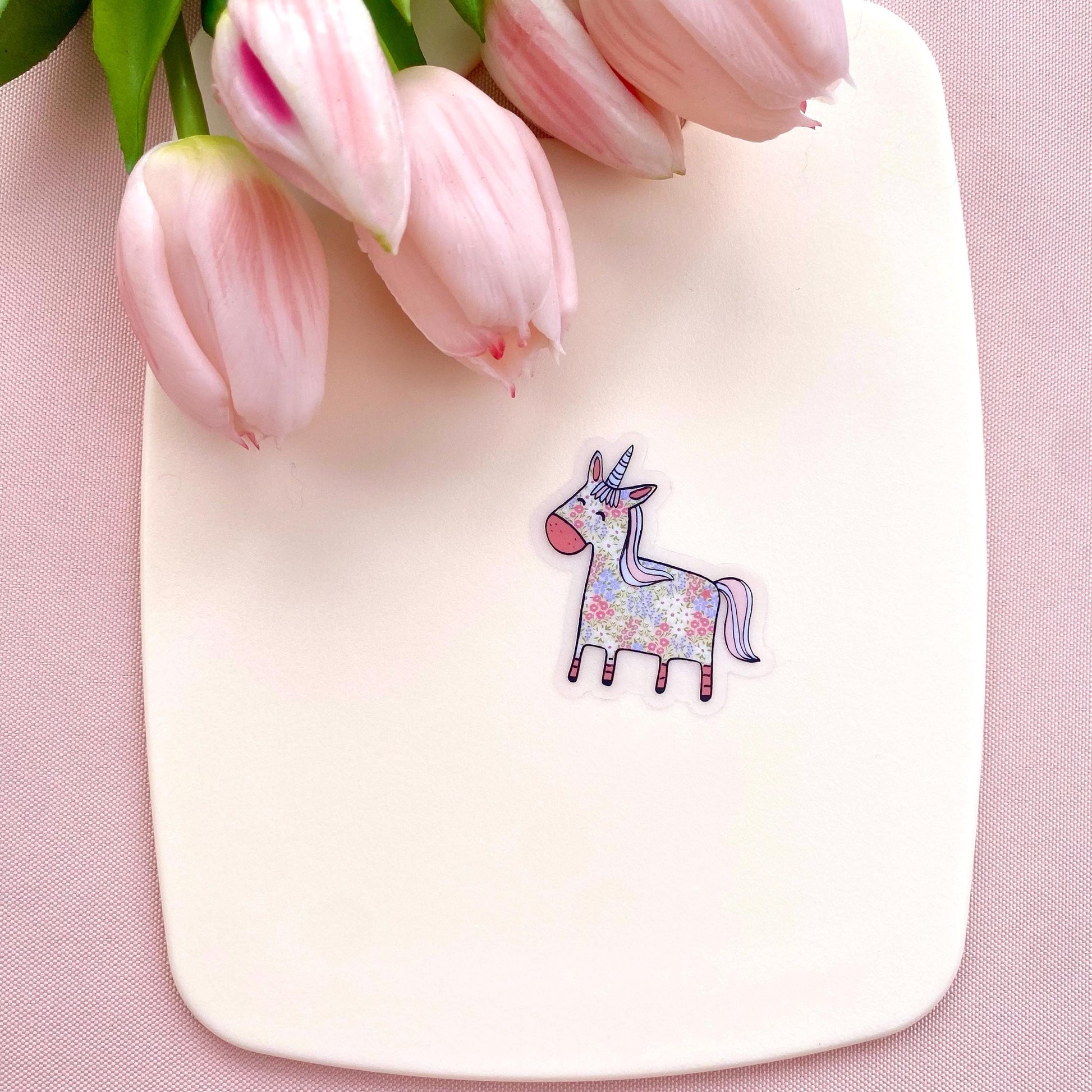 Floral patterned pink and blue unicorn on a transparent vinyl sticker against a white back ground with pink tulips