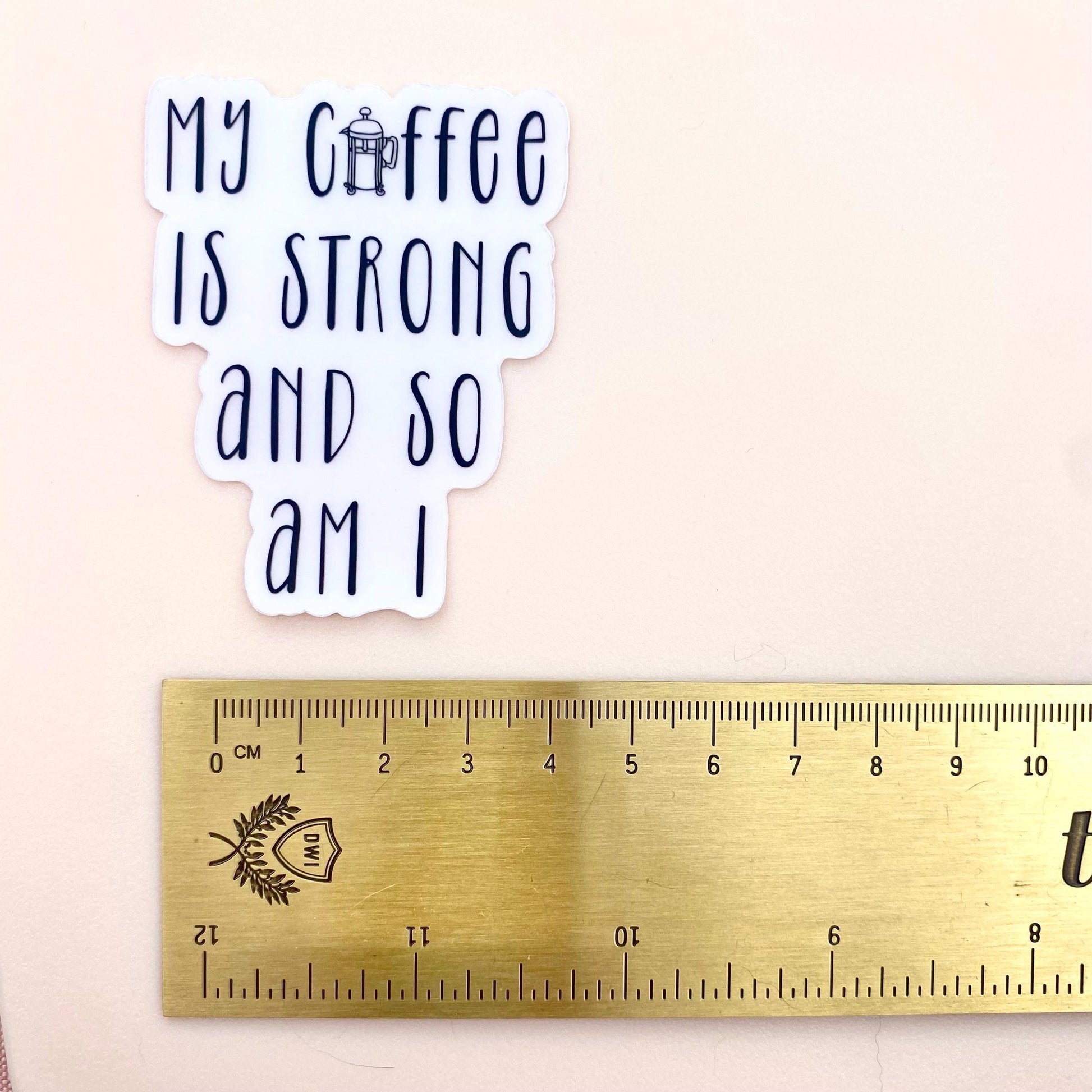 Sticker with 'My coffee is strong and so am I' text next to a ruler for size reference on a beige background