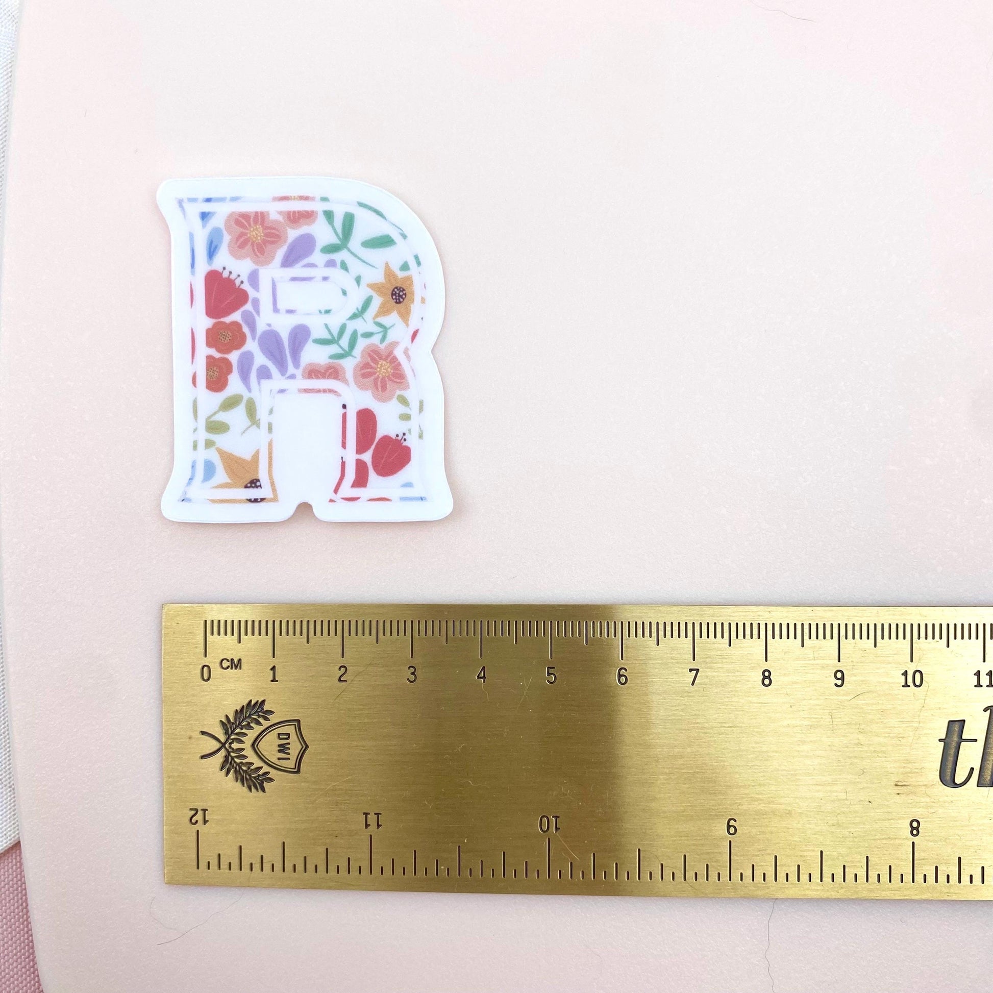 Letter 'R' sticker with floral design on a white background with a gold ruler for scale.