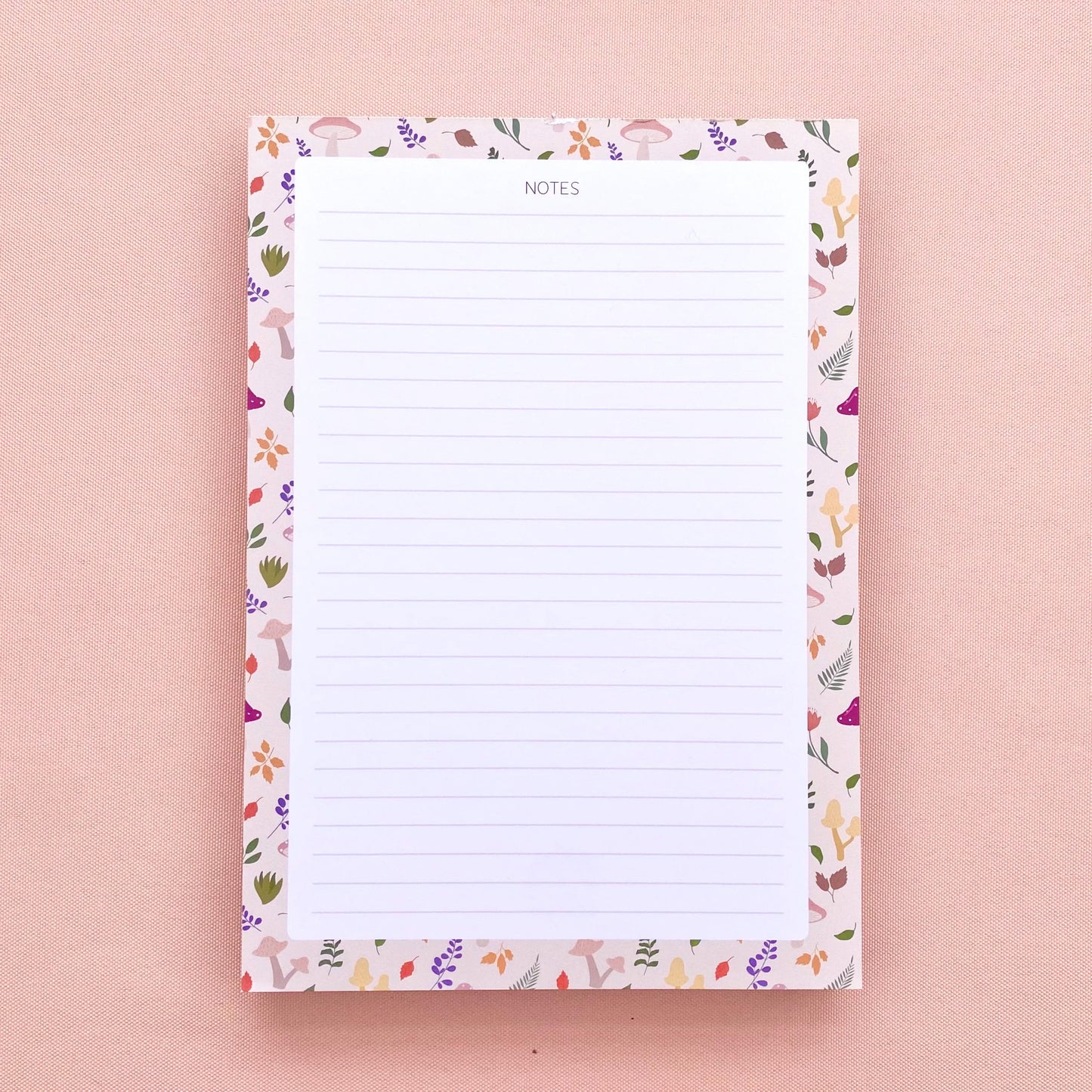 A lined notepad on a pink background, with a beige border patterned with autumn leaves and mushrooms