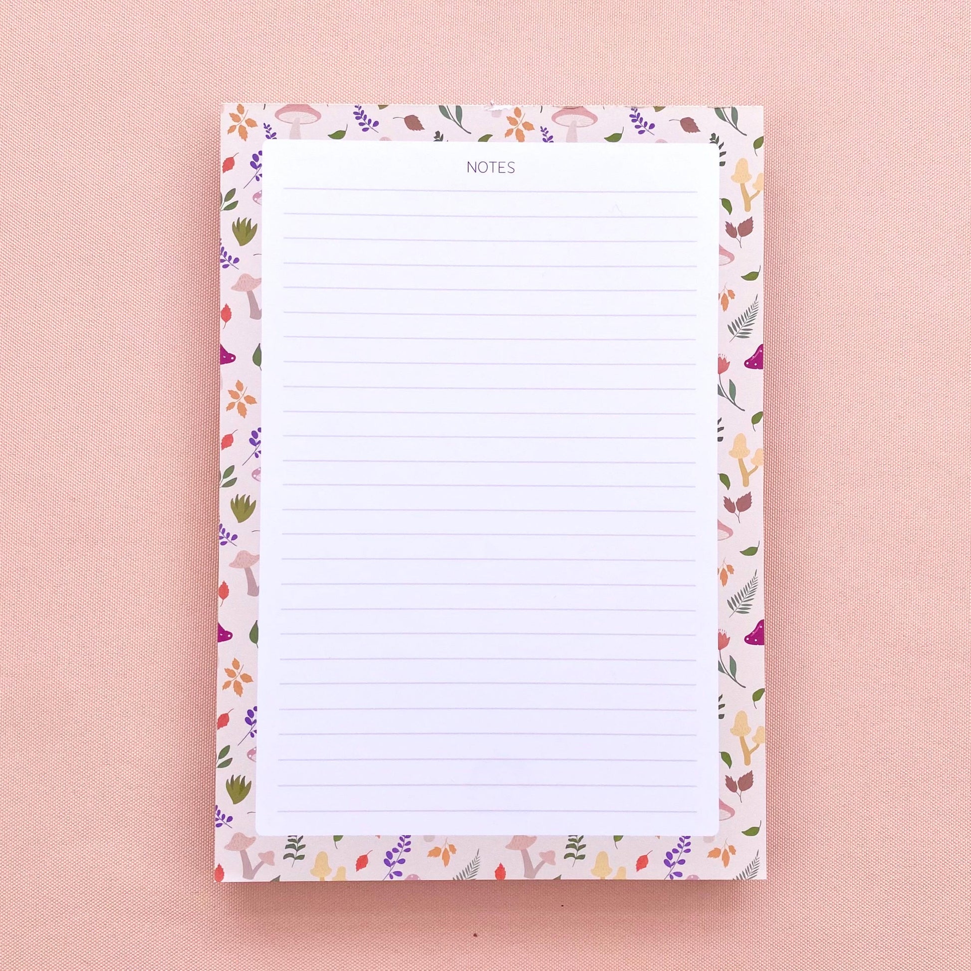 A lined notepad on a pink background, with a beige border patterned with autumn leaves and mushrooms