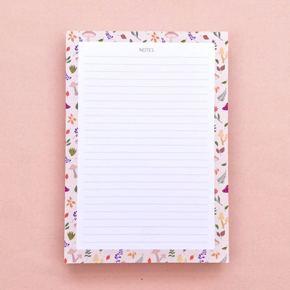 A lined notepad on a pink background, with a beige border patterned with autumn leaves and mushrooms