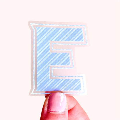 Sticker with a blue and white striped letter 'E' held by a hand on a white background