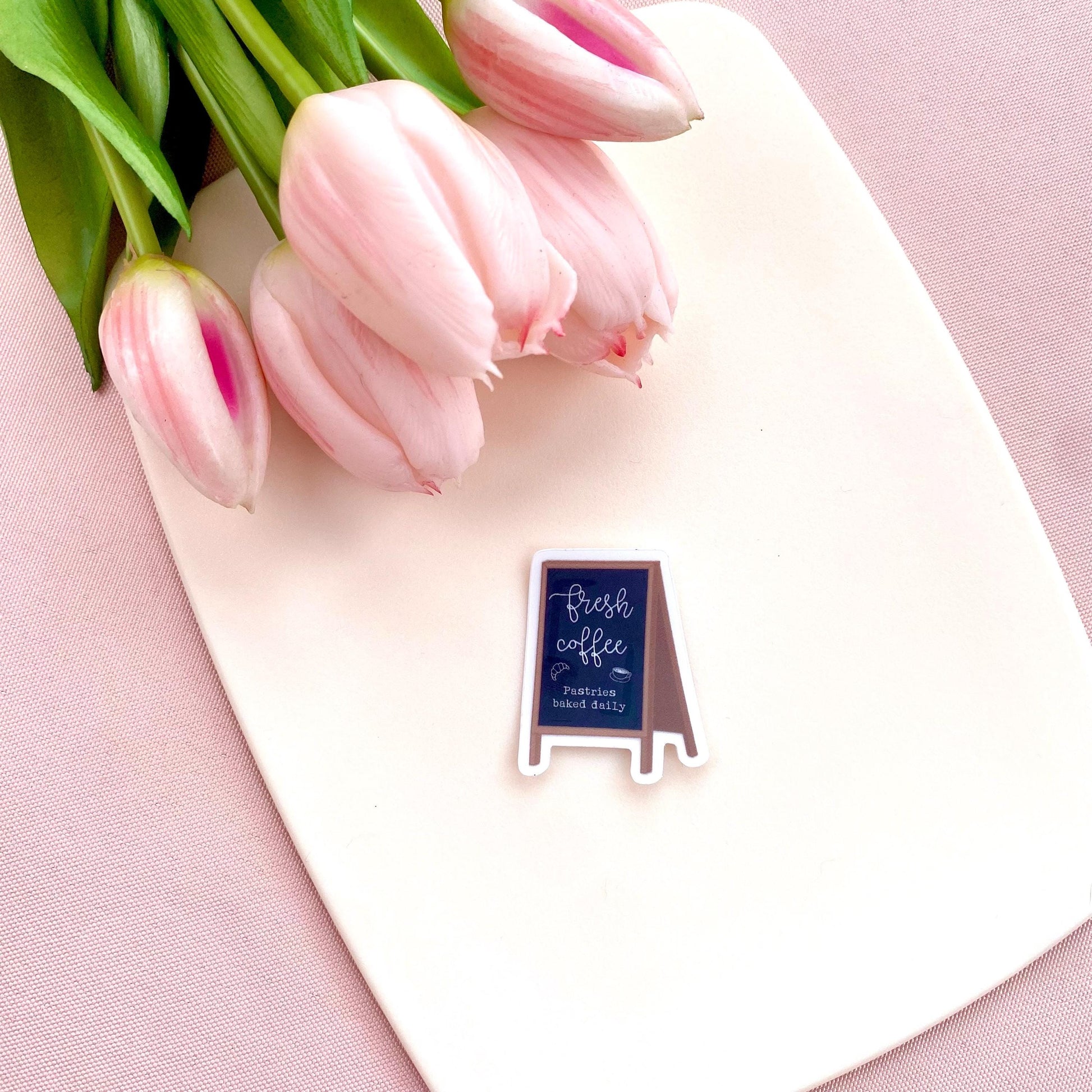 Sticker with a chalkboard sign held by a hand with 'Fresh coffee' and 'Pastries baked daily' text on a white background with pink tulips.