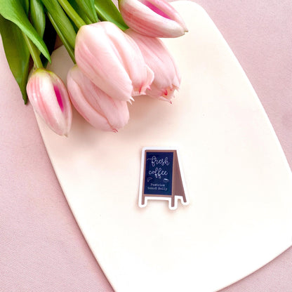 Sticker with a chalkboard sign held by a hand with 'Fresh coffee' and 'Pastries baked daily' text on a white background with pink tulips.