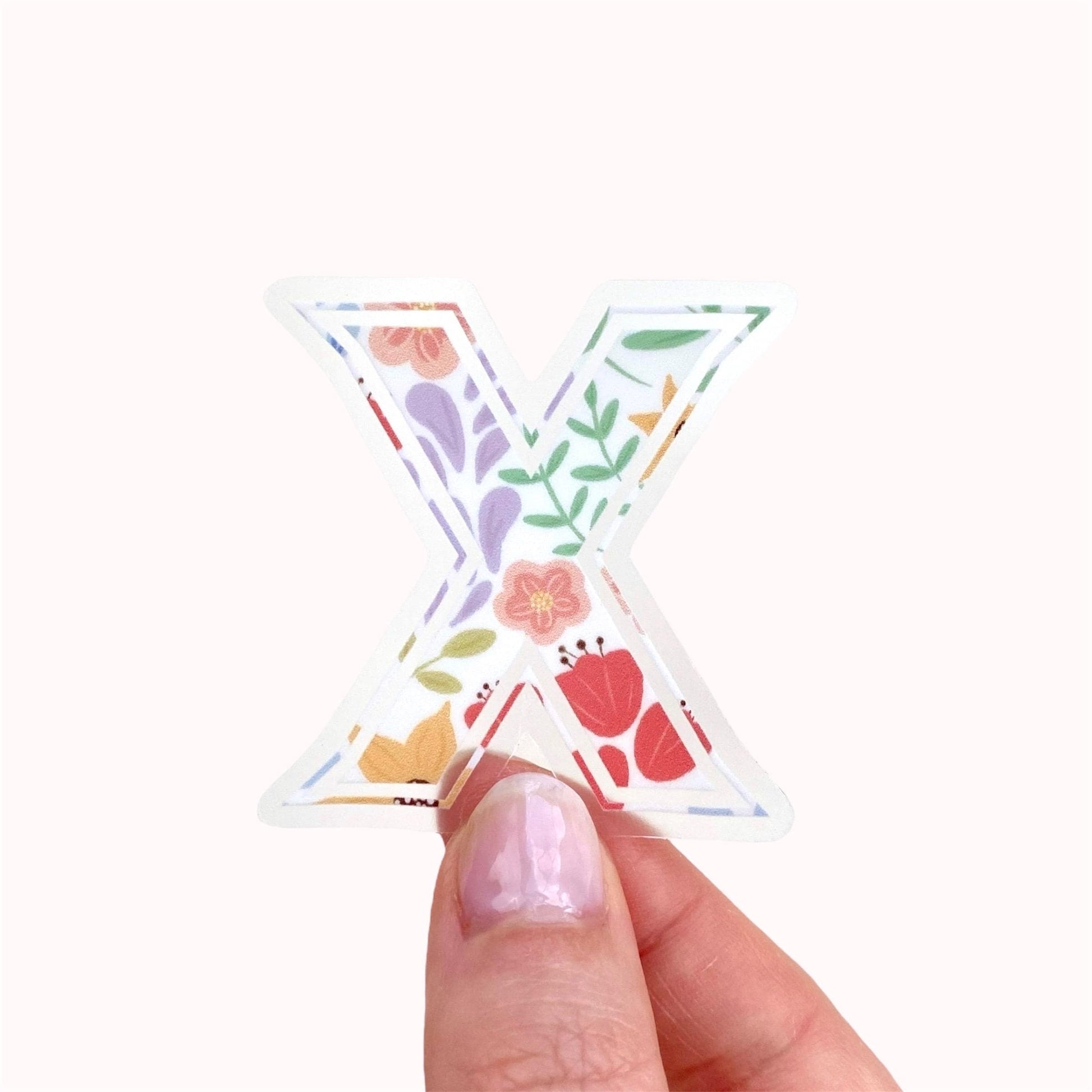 Hand holding a letter 'X' sticker with floral design on a white background