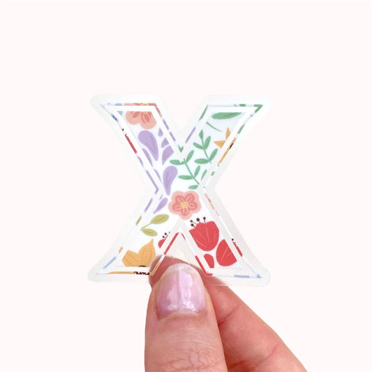 Hand holding a letter 'X' sticker with floral design on a white background
