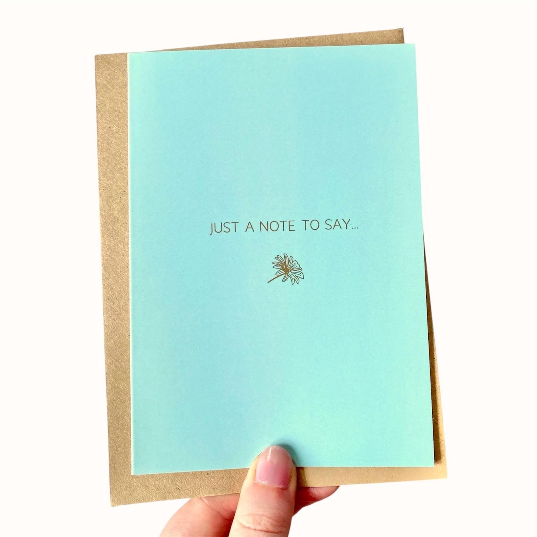 Light blue greeting card with 'Just a note to say...' gold foiled text and dandelion illustration held by a hand.