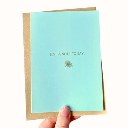 Light blue greeting card with 'Just a note to say...' gold foiled text and dandelion illustration held by a hand.