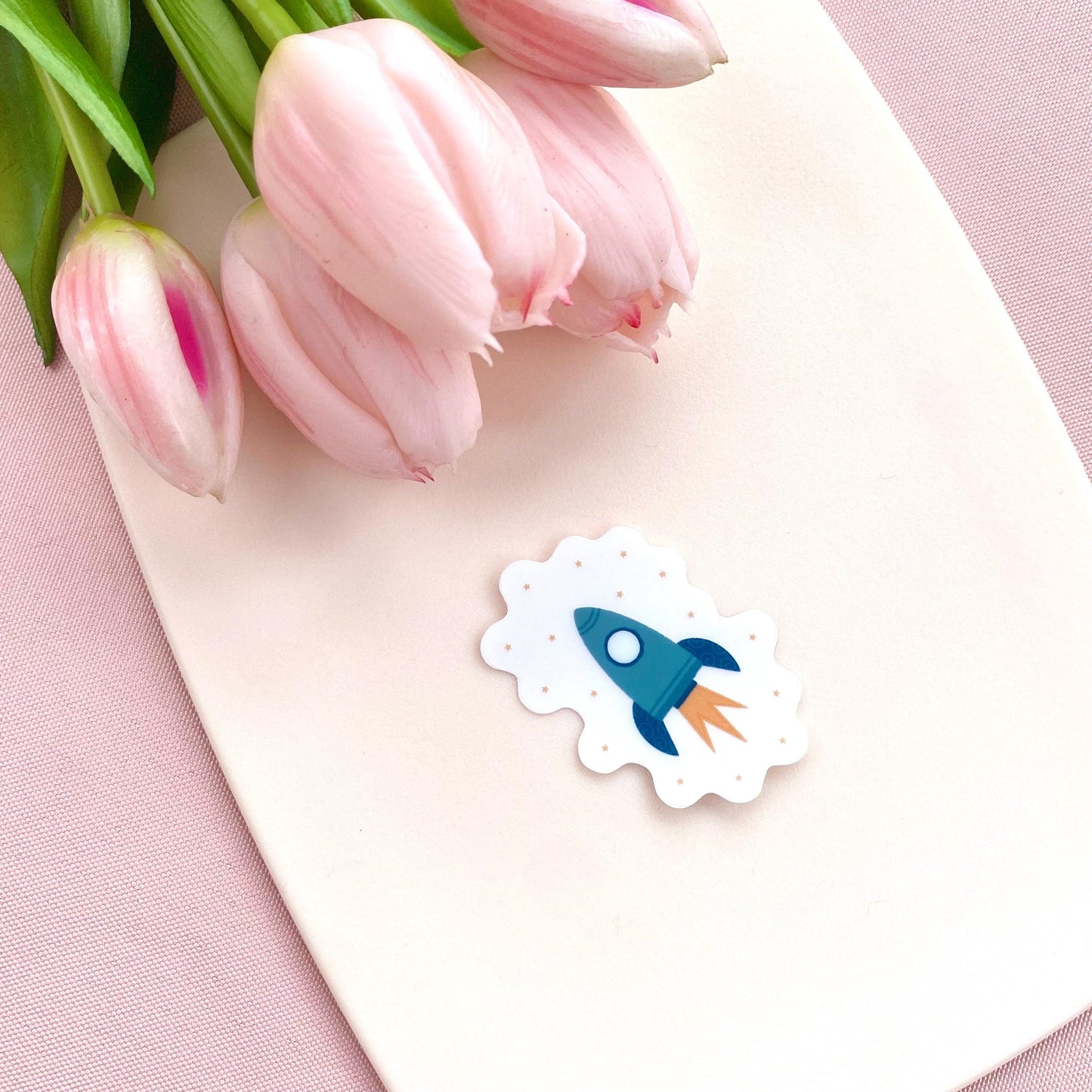 Transparent sticker with teal blue rocket against a cream background with pink tulips