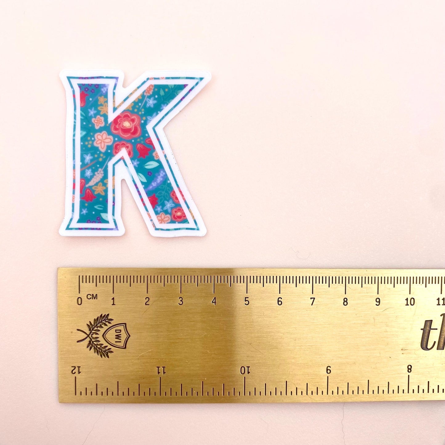 Floral letter 'K' sticker next to a ruler on a beige background